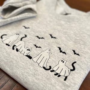 Könnte beinhalten: Ein hellgraues Sweatshirt mit einem gestickten Halloween-Design. Das Design zeigt drei Geisterkatzen und mehrere schwarze Fledermäuse. Das Sweatshirt hat einen Rundhalsausschnitt und lange Ärmel.