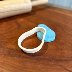 Puede incluir: Un cortador de galletas de plástico blanco con forma de pera, con un cortador de galletas de plástico azul con forma de pera detrás.