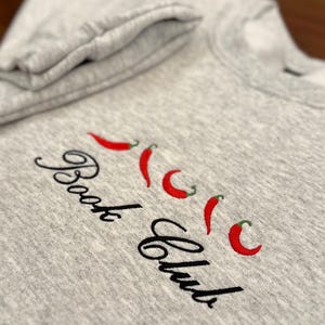 May include: A heather gray sweatshirt with the embroidered text "Book Club" in a cursive font, accompanied by four red chili pepper designs. The sweatshirt's folded sleeve is visible in the upper left corner.