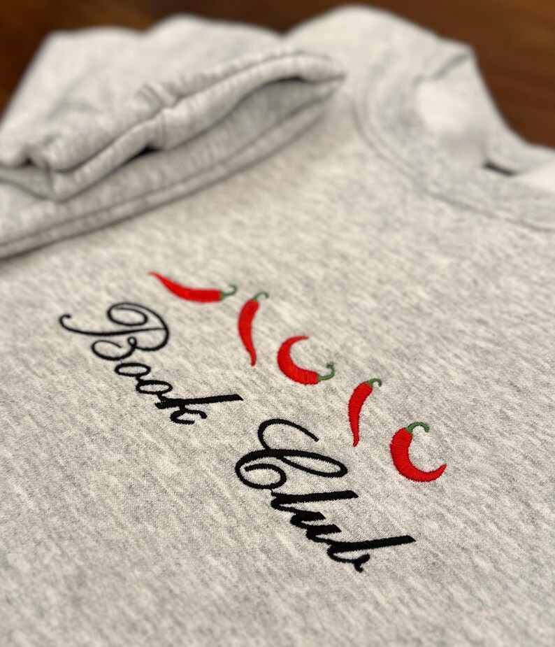 May include: A heather gray sweatshirt with the embroidered text "Book Club" in a cursive font, accompanied by four red chili pepper designs. The sweatshirt's folded sleeve is visible in the upper left corner.