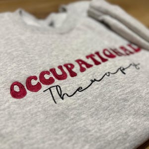 K&ouml;nnte beinhalten: Grauer Sweatshirt mit den Worten "Occupational Therapy" in Rot und Schwarz gestickt.