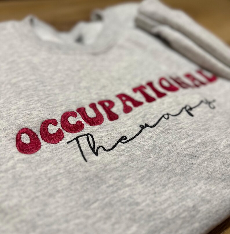 K&ouml;nnte beinhalten: Grauer Sweatshirt mit den Worten "Occupational Therapy" in Rot und Schwarz gestickt.