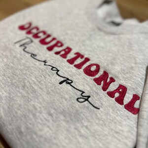 K&ouml;nnte beinhalten: Grauer Pullover mit dem gestickten Text "Occupational Therapy" in Rot.