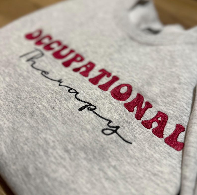 K&ouml;nnte beinhalten: Grauer Pullover mit dem gestickten Text "Occupational Therapy" in Rot.
