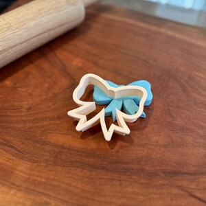 May include: A white plastic cookie cutter shaped like a bow. The cutter is on a wooden surface.