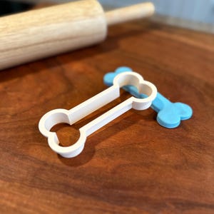 May include: White plastic dog bone cookie cutter with a blue plastic bone cookie cutter in the background. The cookie cutters are on a wooden surface.