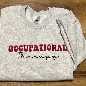 K&ouml;nnte beinhalten: Grauer Sweatshirt mit dem Text "OCCUPATIONAL Therapy" in Rot und Schwarz gestickt.