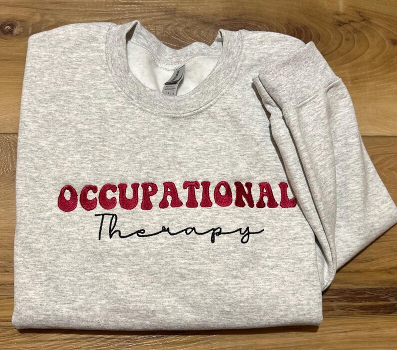 K&ouml;nnte beinhalten: Grauer Sweatshirt mit dem Text "OCCUPATIONAL Therapy" in Rot und Schwarz gestickt.