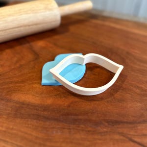 Peut inclure: Un emporte-pièce en plastique blanc en forme de lèvres, sur une surface en bois avec un rouleau à pâtisserie en arrière-plan.