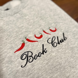 May include: A light gray sweatshirt with embroidered red chili peppers and the words "Book Club" in black script. The design features four chili peppers with green stems.
