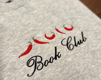 Puede incluir: Sudadera gris claro con chiles rojos bordados y las palabras "Book Club" en escritura negra. El diseño presenta cuatro chiles con tallos verdes.