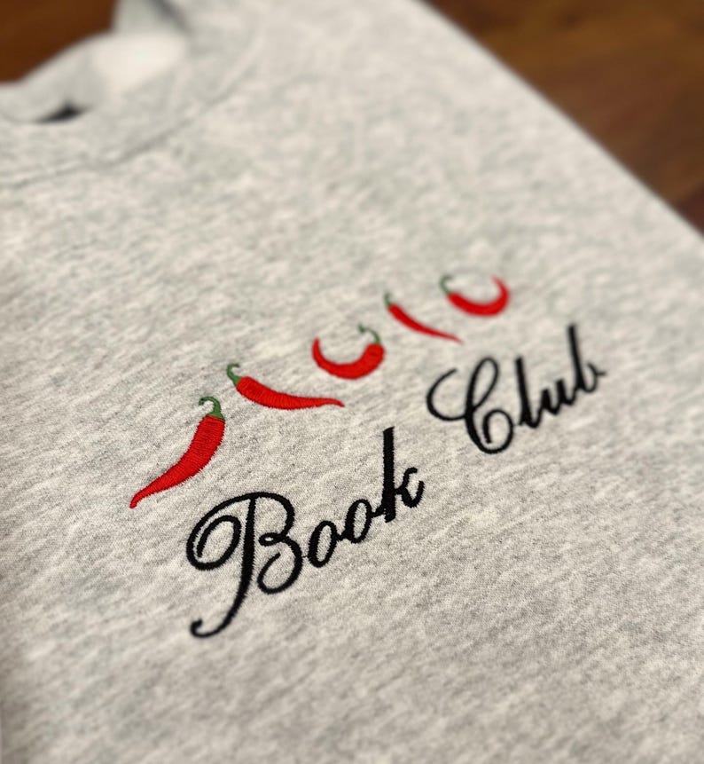 May include: A light gray sweatshirt with embroidered red chili peppers and the words "Book Club" in black script. The design features four chili peppers with green stems.