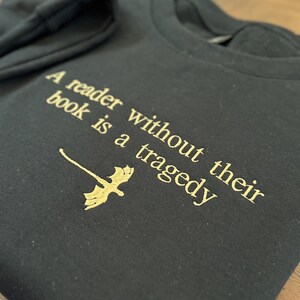 Op de afbeelding: Zwart sweatshirt met gouden tekst die luidt: "Een lezer zonder zijn boek is een tragedie." Een gouden draakafbeelding staat onder de tekst. Het sweatshirt is op een houten oppervlak gevouwen.