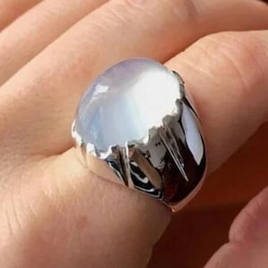 Pode incluir: Um anel de prata com uma grande pedra preciosa oval azul clara. O anel tem um design texturizado e irregular em torno da pedra preciosa. O anel está no dedo de uma pessoa.