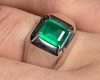 May include: A silver ring featuring a large, square-cut emerald gemstone. The emerald has a vibrant green color and is set in a polished silver band. The ring is shown on a person's finger, highlighting its design and size.