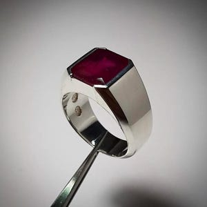 Puede incluir: Un anillo de plata con una gran gema rectangular de color rojo intenso. El anillo tiene un diseño pulido y moderno con una banda gruesa y un engaste biselado para el rubí. Dos pequeñas piedras redondas de acento son visibles en la banda.
