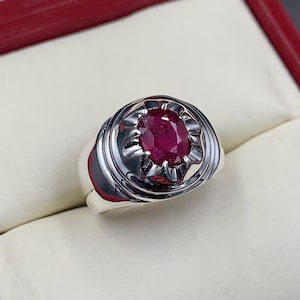 May include: A silver ring with a red oval ruby gemstone set in a decorative bezel.