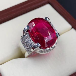 May include: A silver ring with a large, oval-shaped, red gemstone surrounded by smaller, clear gemstones.