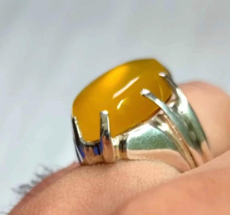 May include: A silver ring with a yellow oval gemstone set in a prong setting. The ring has a decorative band with a blue accent.
