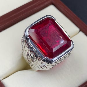 May include: A silver ring with an ornate design and a large, deep red, emerald-cut gemstone.