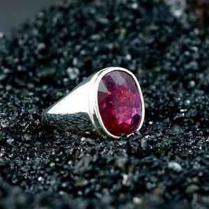 Puede incluir: Un anillo de plata con una gran gema de rubí de corte ovalado. El rubí es de color rojo intenso y está engastado en una banda de plata pulida. El anillo se muestra sobre una superficie oscura y texturizada.