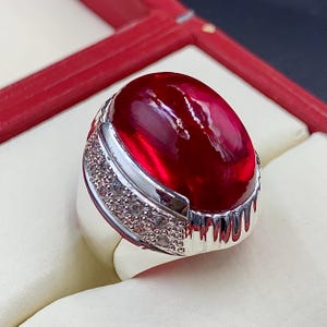 May include: A silver ring with a large, oval, deep red gemstone. The ring has a silver band with a row of small, clear stones. The polished gemstone reflects light. The ring is presented in a red box.