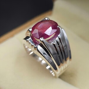 May include: A silver ring featuring a large, oval-shaped ruby gemstone. The ring has a thick band with a textured design.