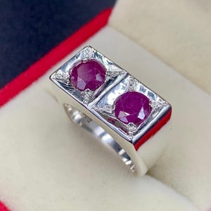 May include: A silver ring with two oval, deep red rubies set next to each other. The rubies are framed by small, clear accent stones. The ring has a rectangular face and a polished finish, set against a red and cream background.