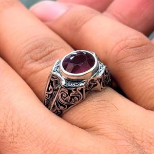 Puede incluir: Un anillo de plata con un diseño adornado y una gran piedra preciosa de color rojo oscuro de forma ovalada.