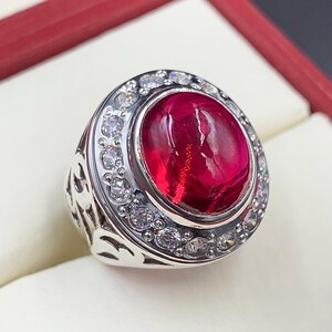 May include: A silver ring with a large, oval-shaped, red gemstone surrounded by a halo of smaller, clear gemstones.