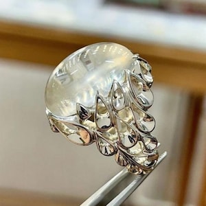 Pode incluir: Um anel de prata com uma grande gema oval transparente. A banda do anel tem um design decorativo vazado com elementos em forma de gota. A gema é mantida no lugar por uma cravação de prata. O anel é segurado por uma pinça.