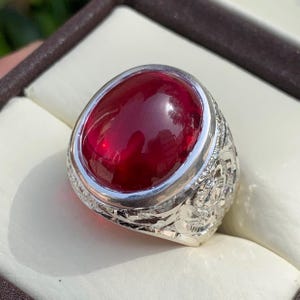 May include: A silver ring with a large, oval-shaped, red gemstone set in a bezel setting. The band of the ring is intricately engraved with a floral design.