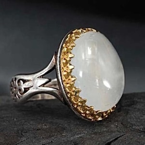 Pode incluir: Anel de prata com uma grande pedra preciosa branca oval. A pedra é engastada em uma moldura dourada com borda ondulada. A banda do anel tem um design ornamentado e vazado.