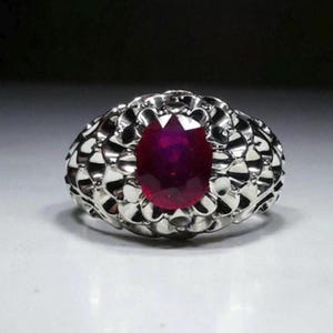 Könnte beinhalten: Ein silberner Ring mit einem großen, oval geschliffenen Rubin. Das Band des Rings weist ein detailliertes, strukturiertes Design auf. Der Rubin hat eine tiefrote Farbe, und der Ring ist vor einem neutralen Hintergrund platziert.
