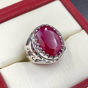 May include: A silver ring featuring an intricate design and a large, oval ruby gemstone.