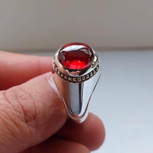 May include: A silver ring with a red oval gemstone set in a bezel setting. The ring has a beaded edge around the gemstone.