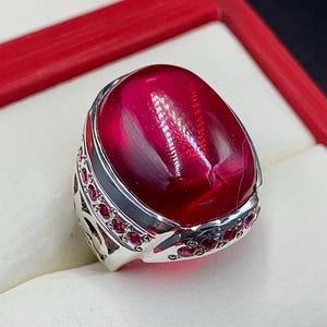 May include: A silver ring with a large, oval-shaped, cabochon ruby gemstone. The ring is set with smaller ruby stones around the gemstone.