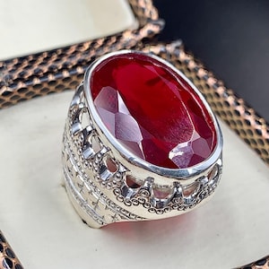 May include: A silver ring featuring an oval red gemstone set in an intricate, ornate design.