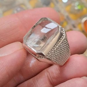 Pode incluir: Um anel de pedra preciosa transparente e retangular com uma banda prateada. A pedra preciosa tem um interior ligeiramente nublado. A banda apresenta um design geométrico texturizado. O anel é segurado em uma mão.