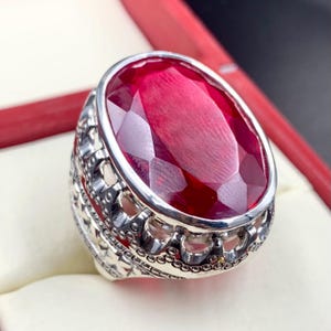 Può includere: Un anello d'argento con un designo ornato e una grande pietra rossa di forma ovale.