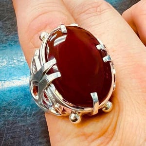 Puede incluir: Un anillo de plata con una gran piedra de cornalina de forma ovalada. El anillo tiene un diseño detallado e intrincado con una banda de plata.
