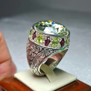 May include: A silver ring featuring a large, oval, light green gemstone. The ring is embellished with smaller green and red gemstones. The band has detailed floral carvings. The ring is displayed on a small stand.