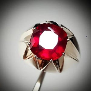 May include: A gold-toned ring featuring a large, faceted, deep red gemstone. The gemstone is the focal point, with a bright, reflective center. The ring's band has a classic, simple design.