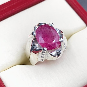 May include: A silver ring with a large, oval-shaped, pink ruby gemstone set in a prong setting. The ring has a detailed, ornate design.