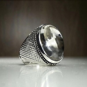 Pode incluir: Um anel de prata com uma pedra grande, oval e transparente. A banda apresenta um padrão geométrico detalhado e uma borda de pequenas contas. O anel é colocado sobre um fundo neutro, destacando o seu acabamento polido.