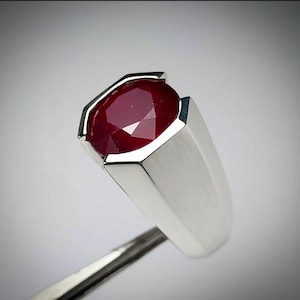 May include: A silver ring with a large, oval-cut ruby gemstone. The ruby is a deep red colour and is set in a modern, geometric silver band. The ring is held by a metal tool.