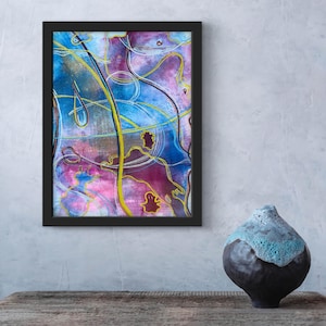 Puede incluir: Impresión de arte abstracto enmarcada en negro con colores azul, morado y amarillo. Un jarrón oscuro con una parte superior texturizada azul claro descansa sobre una mesa de madera. La pared es gris.