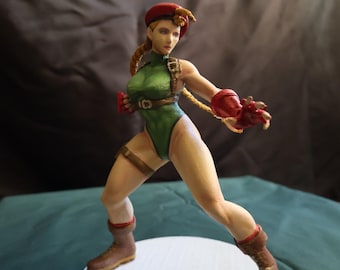 May include: A figurine of a female character in a fighting stance. She wears a green bodysuit, red gloves, and a red beret. Her boots are brown, and she has blonde hair. The figure is on a white circular base.