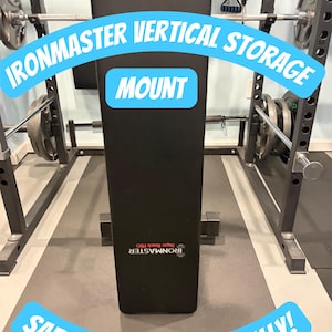 May include: Black weight bench with the text "IRONMASTER VERTICAL STORAGE MOUNT SAFELY STORE IT VERTICALLY!" in a home gym setting. The bench is positioned vertically, with weight plates and a barbell visible in the background.