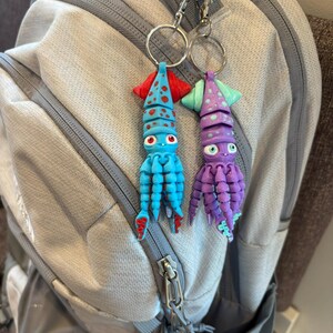 May include: Two whimsical squid keychains. One is blue with red accents, the other is purple with green accents. Both have large eyes and are attached to silver keyrings. They are hanging in front of a gray backpack.
